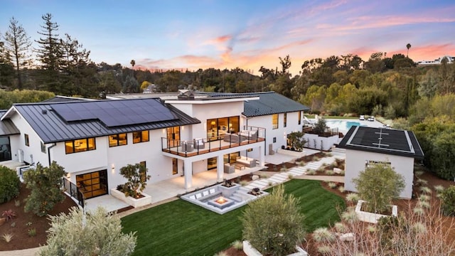 back house at dusk featuring a balcony, an outdoor living space with a fire pit, a patio, and a lawn