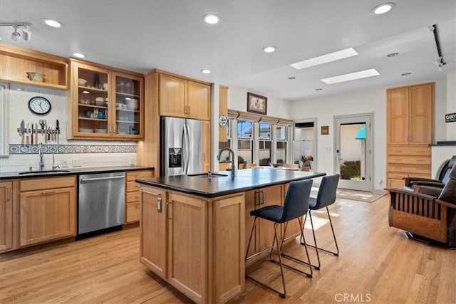 kitchen featuring dark countertops, appliances with stainless steel finishes, glass insert cabinets, a kitchen island with sink, and a sink