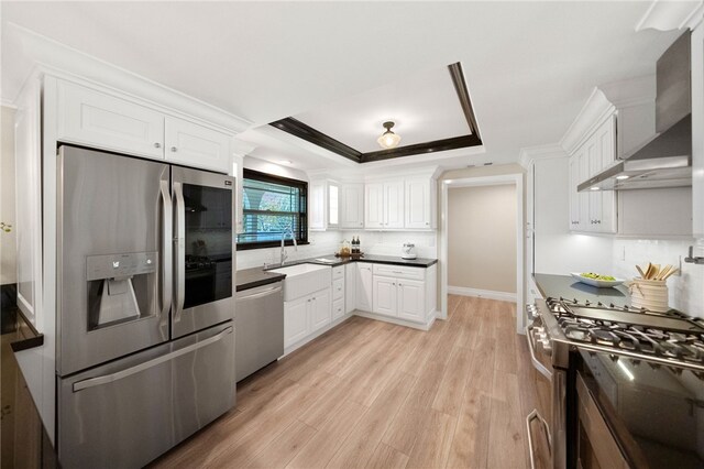 kitchen featuring wall chimney range hood, sink, appliances with stainless steel finishes, white cabinetry, and a raised ceiling