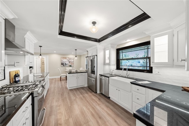 kitchen featuring appliances with stainless steel finishes, white cabinets, hanging light fixtures, a tray ceiling, and wall chimney range hood