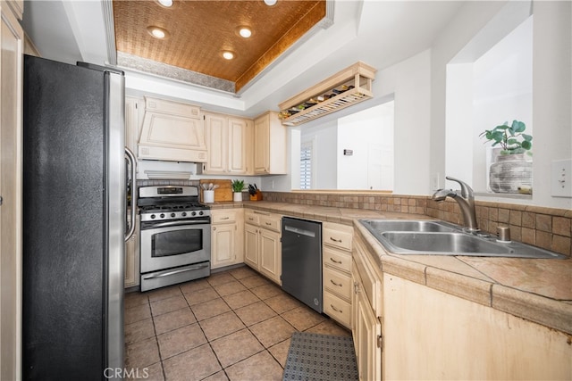 kitchen with a raised ceiling, stainless steel appliances, cream cabinetry, premium range hood, and a sink