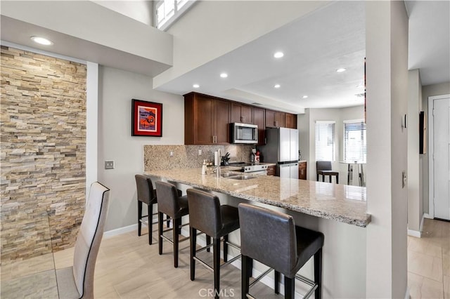 kitchen with backsplash, stainless steel appliances, light stone counters, a kitchen bar, and kitchen peninsula