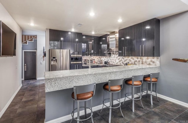 kitchen with a breakfast bar area, decorative backsplash, kitchen peninsula, stainless steel appliances, and wall chimney exhaust hood