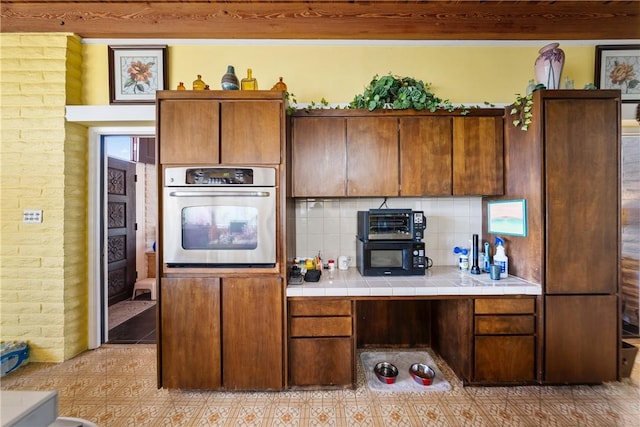 kitchen featuring stainless steel oven, brick wall, tile counters, and backsplash