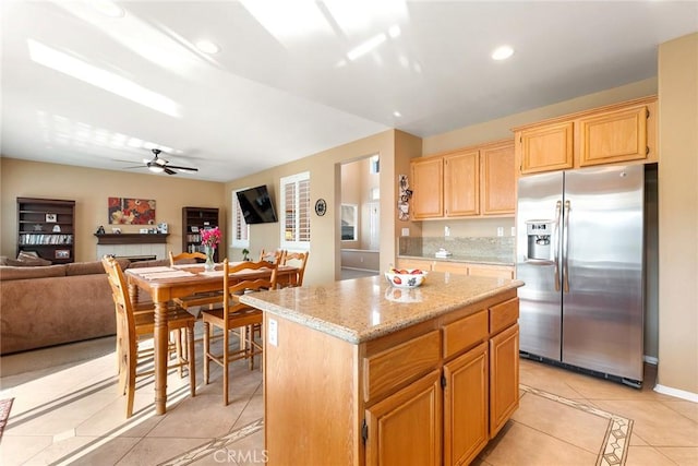 kitchen with light tile patterned flooring, light stone counters, stainless steel fridge, a kitchen island, and ceiling fan