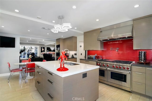 kitchen with pendant lighting, wall chimney range hood, a kitchen island with sink, backsplash, and stainless steel appliances