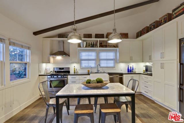 kitchen with pendant lighting, appliances with stainless steel finishes, white cabinetry, lofted ceiling with beams, and wall chimney exhaust hood