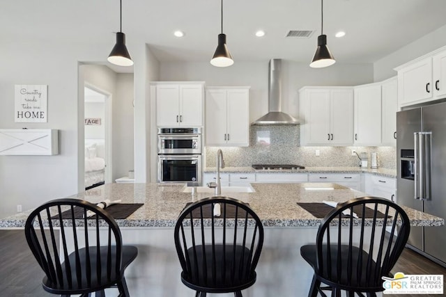 kitchen with white cabinetry, stainless steel appliances, a center island with sink, and wall chimney range hood