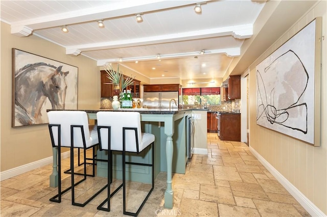 kitchen featuring a breakfast bar, stainless steel built in refrigerator, tasteful backsplash, an island with sink, and beamed ceiling