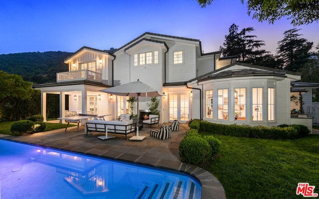 back house at dusk featuring an outdoor living space, a balcony, a lawn, and a patio area