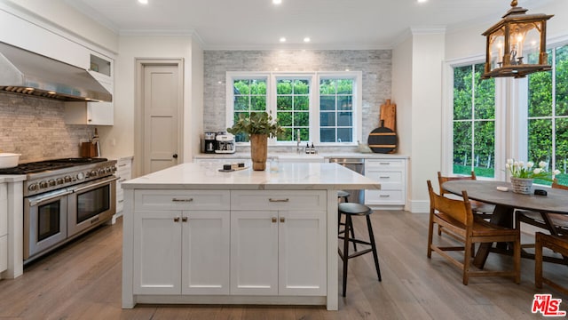 kitchen with wall chimney range hood, white cabinetry, stainless steel appliances, light stone counters, and ornamental molding