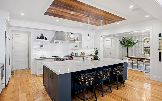 kitchen with white cabinetry, wall chimney exhaust hood, light stone countertops, and a center island with sink