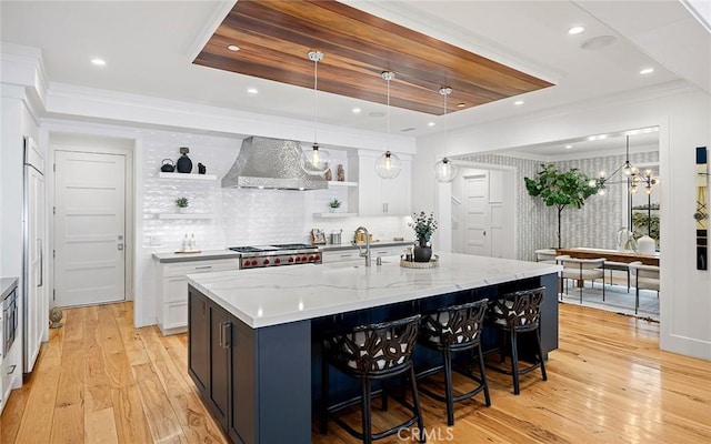 kitchen with open shelves, light wood-style flooring, a sink, wall chimney range hood, and a large island with sink