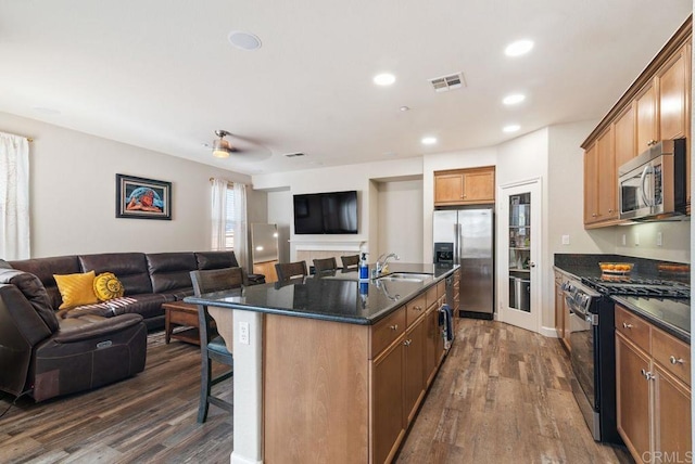 kitchen with open floor plan, stainless steel appliances, dark countertops, and a kitchen island with sink