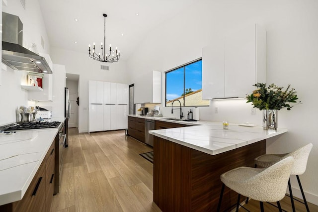 kitchen featuring wall chimney exhaust hood, decorative light fixtures, stainless steel appliances, light stone countertops, and white cabinets