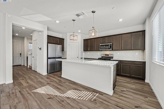 kitchen with hardwood / wood-style flooring, appliances with stainless steel finishes, hanging light fixtures, backsplash, and a center island with sink