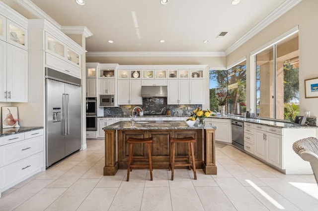 kitchen featuring a kitchen breakfast bar, built in appliances, white cabinets, a center island with sink, and dark stone counters