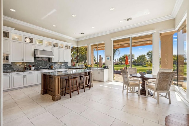 kitchen featuring stainless steel microwave, range hood, white cabinets, dark stone counters, and a center island with sink