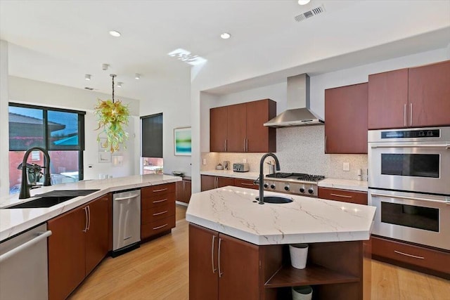 kitchen with sink, appliances with stainless steel finishes, tasteful backsplash, an island with sink, and wall chimney exhaust hood