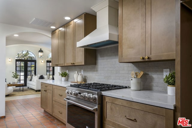 kitchen with wall chimney range hood, stainless steel range, light stone counters, and decorative backsplash