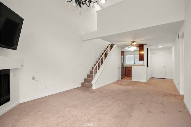 unfurnished living room featuring stairs, a fireplace, light carpet, baseboards, and ceiling fan with notable chandelier