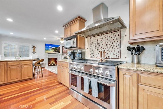 kitchen featuring wall chimney range hood, appliances with stainless steel finishes, light brown cabinets, decorative backsplash, and light wood-type flooring