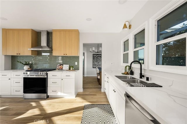 kitchen with light brown cabinets, a sink, white cabinetry, wall chimney range hood, and appliances with stainless steel finishes