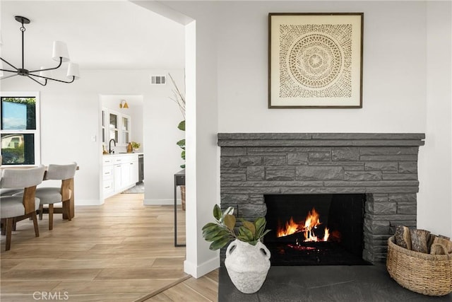 living area featuring a stone fireplace, light wood-style flooring, a notable chandelier, visible vents, and baseboards