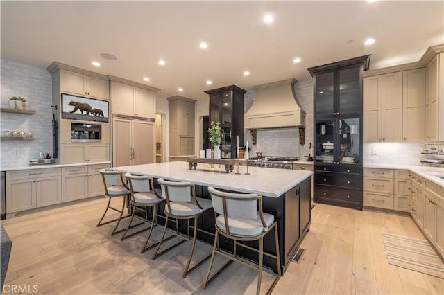 kitchen featuring light countertops, a kitchen island with sink, light wood-type flooring, premium range hood, and a kitchen breakfast bar