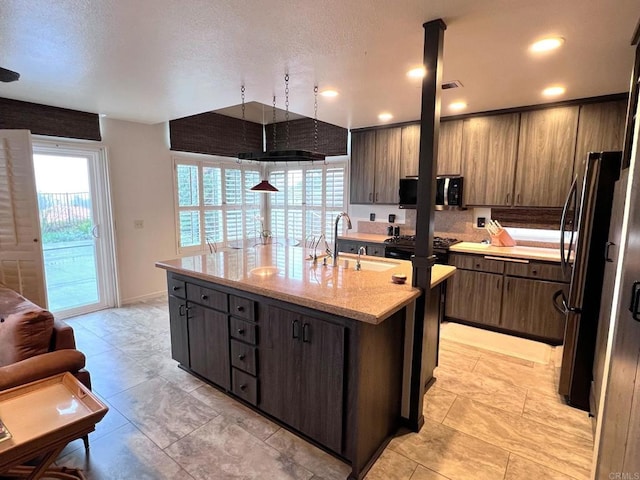 kitchen with dark brown cabinetry, sink, light stone counters, stainless steel appliances, and a kitchen island with sink