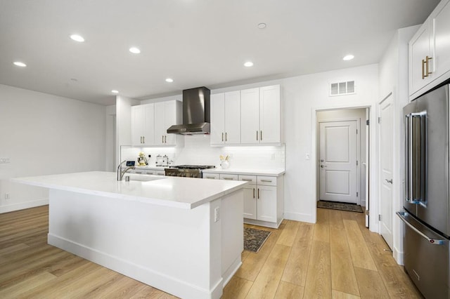 kitchen featuring wall chimney range hood, a kitchen island with sink, white cabinetry, tasteful backsplash, and high end refrigerator