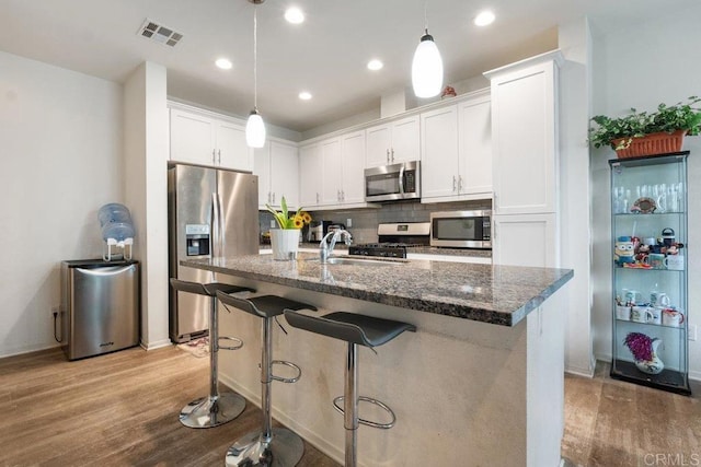 kitchen featuring light hardwood / wood-style flooring, appliances with stainless steel finishes, an island with sink, dark stone counters, and white cabinets