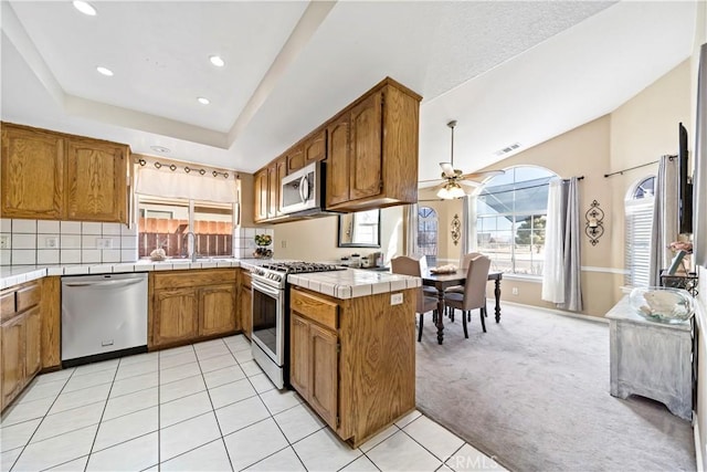 kitchen with appliances with stainless steel finishes, kitchen peninsula, tile counters, a tray ceiling, and light carpet