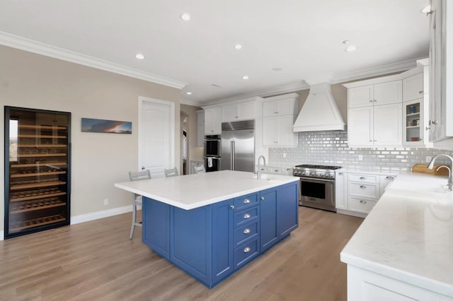 kitchen with high end appliances, custom range hood, a kitchen island with sink, blue cabinetry, and white cabinetry