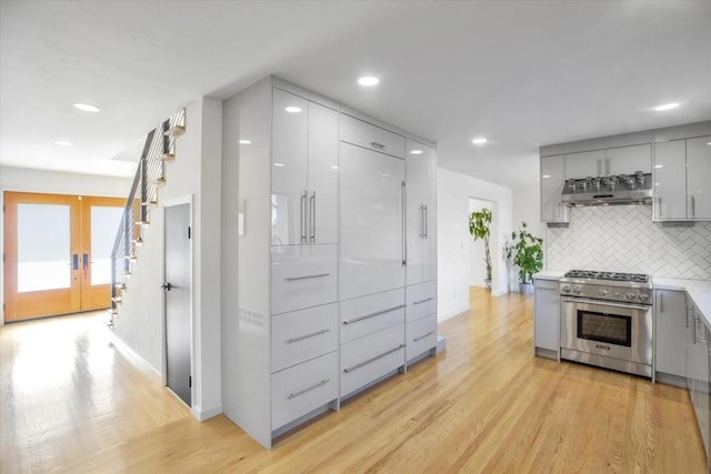 kitchen featuring light wood-type flooring, gray cabinets, stainless steel stove, range hood, and decorative backsplash