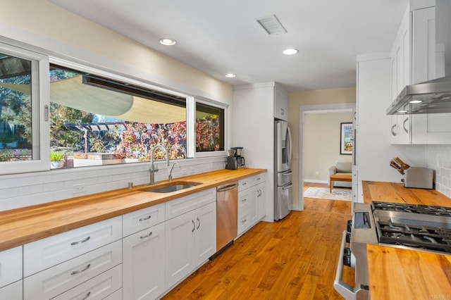 kitchen with sink, white cabinets, wooden counters, stainless steel appliances, and wall chimney exhaust hood