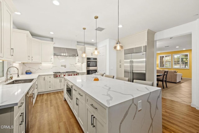 kitchen featuring sink, built in appliances, a kitchen island, wall chimney range hood, and white cabinets