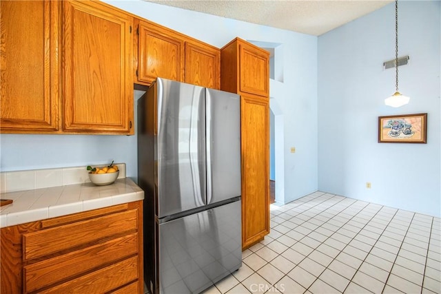 kitchen featuring tile counters, brown cabinetry, freestanding refrigerator, and decorative light fixtures