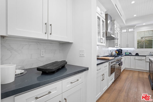 kitchen featuring wall chimney range hood, backsplash, ornamental molding, light hardwood / wood-style floors, and white cabinets