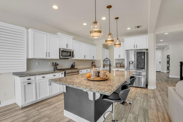 kitchen featuring an island with sink, pendant lighting, stainless steel appliances, light stone countertops, and white cabinets