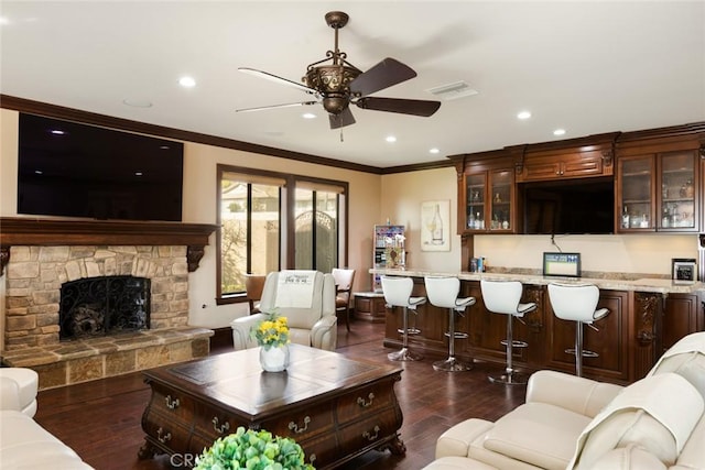 living room featuring a stone fireplace, indoor bar, dark hardwood / wood-style flooring, ornamental molding, and ceiling fan