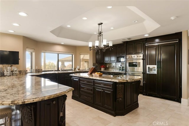 kitchen with hanging light fixtures, built in appliances, dark brown cabinetry, and a tray ceiling