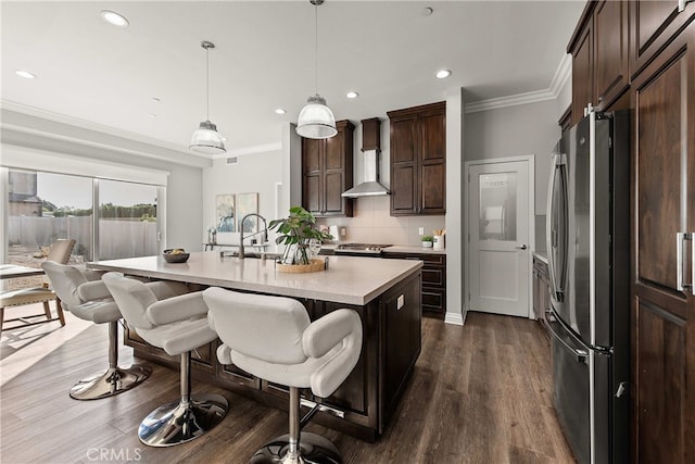 kitchen featuring wall chimney exhaust hood, a center island with sink, ornamental molding, stainless steel fridge, and pendant lighting