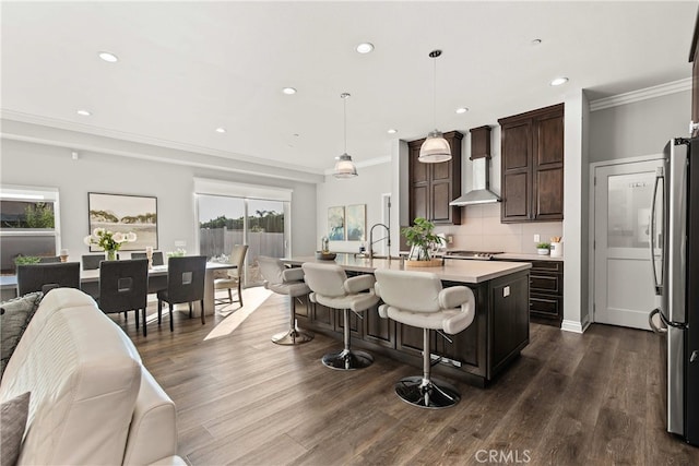 kitchen with stainless steel refrigerator, decorative light fixtures, an island with sink, backsplash, and wall chimney range hood