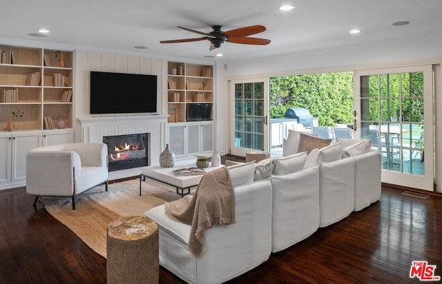 living room featuring crown molding, dark wood-type flooring, ceiling fan, a fireplace, and built in shelves