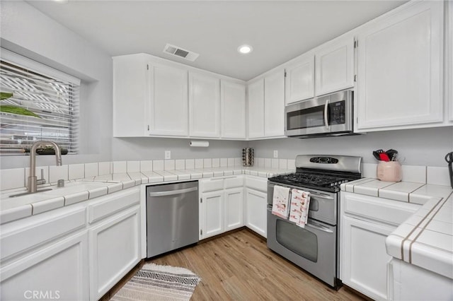 kitchen with appliances with stainless steel finishes, tile countertops, white cabinetry, sink, and light hardwood / wood-style flooring