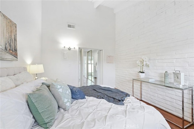 bedroom featuring beamed ceiling, a towering ceiling, brick wall, and wood-type flooring