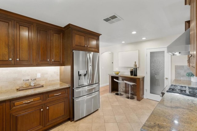 kitchen with light stone countertops, wall chimney range hood, decorative backsplash, and stainless steel fridge with ice dispenser