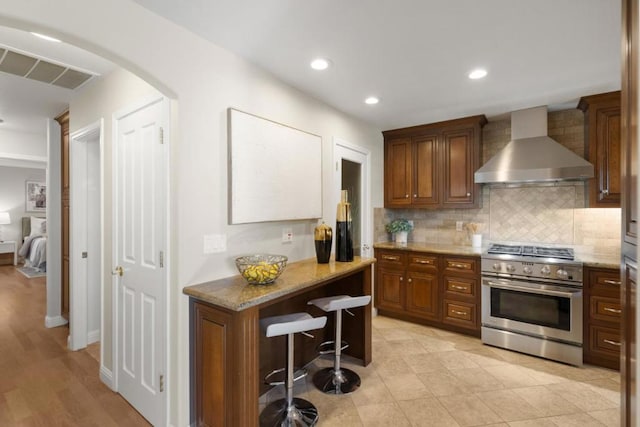 kitchen featuring wall chimney exhaust hood, a breakfast bar, tasteful backsplash, light stone counters, and high end stove