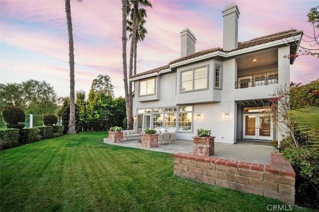 back house at dusk with french doors, a balcony, a patio, and a lawn